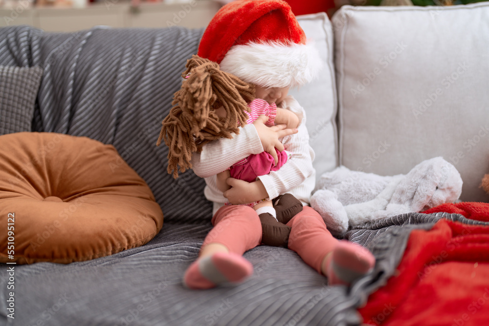 Adorable caucasian girl hugging baby doll sitting on sofa by christmas decoration at home
