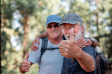 Blurred couple of old active men with hat and backpack have fun in mountain hike in the woods looking at camera with thumb up. Adventure is ageless. Retired seniors and healthy lifestyle concept