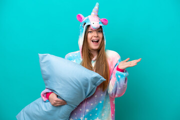 Young caucasian girl with unicorn pajamas holding pillow isolated on blue background with shocked facial expression