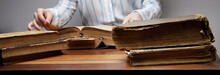  The Hands Of An Invisible Person Leaf Through The Pages Of An Old Book Lying On A Wooden Table In A University Or School Library.vintage Paper Texture. Selective Focus