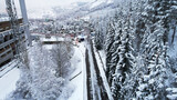 Fototapeta Do pokoju - Snowy fairy-tale road in a mountain forest. Christmas or New Year has come. Coniferous trees in the snow. There is a green bus, people are walking. Light fog. The view from the drone. Medeo, Almaty