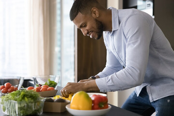 Happy handsome young African chef guy cooking vegan meal, chopping fresh vegetables for salad at kitchen table, keeping healthy lifestyle diet. Blogger man cutting organic ingredients