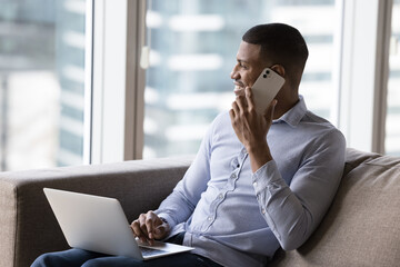 Wall Mural - Happy dreamy African digital gadget user man talking on cell, enjoying phone call, looking at window, smiling, keeping laptop on lap. Young businessman working from home, hotel room