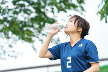 サッカー観戦しながら熱中症対策・暑さ対策のため水を飲むサッカーファン・サポーターのアジア人女性
