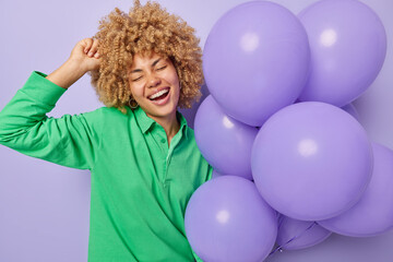 Wall Mural - Festive occasion partying concept. Cheerful curly haired woman laughs gladfully holds bunch of inflated balloons enjoys birthday celebration smiles broadly keeps eyes closed isolated over purple wall