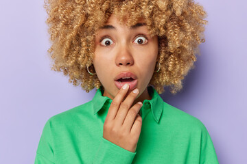 Wall Mural - Cropped shot of amazed curly haired young woman stares with widely opened eyes and mouth reacts to something shocking wears green jumper isolated over purple background. Human reactions concept