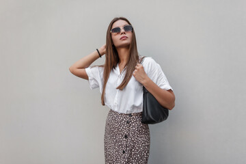 Wall Mural - Fashionable beautiful summer woman model with vintage sunglasses in fashion clothes with a white shirt and a skirt with a trendy black bag stands near a gray building in the city