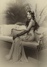 Portrait Flapper Woman In Twenties Old Style Sitting On Sofa, Couch. Long Evening Retro Dress Ostrich Feather Boa Cold Wave Hairstyle. Art Sepia Black And White Vintage Photo. Roar 1920s Fashion Model