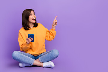 Sticker - Full body length photo of young excited nice lady sitting finger pointing surprised mockup offer online shopping hold phone isolated on purple color background