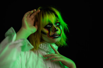 Wall Mural - Portrait of woman with yellow hair and evil clown face art posing on black background, green side lighting