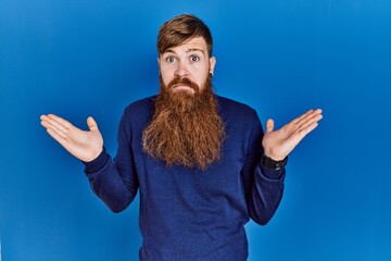 Canvas Print - Redhead man with long beard wearing casual blue sweater over blue background clueless and confused expression with arms and hands raised. doubt concept.