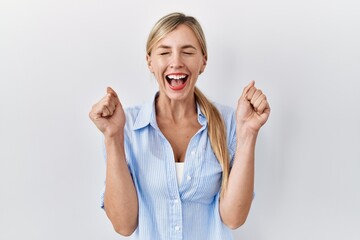 Sticker - Beautiful blonde woman standing over white background excited for success with arms raised and eyes closed celebrating victory smiling. winner concept.