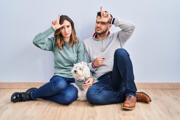 Canvas Print - Young hispanic couple sitting on the floor with dog making fun of people with fingers on forehead doing loser gesture mocking and insulting.