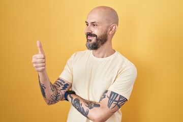 Wall Mural - Hispanic man with tattoos standing over yellow background looking proud, smiling doing thumbs up gesture to the side