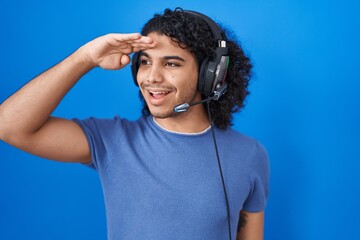 Hispanic man with curly hair listening to music using headphones very happy and smiling looking far away with hand over head. searching concept.