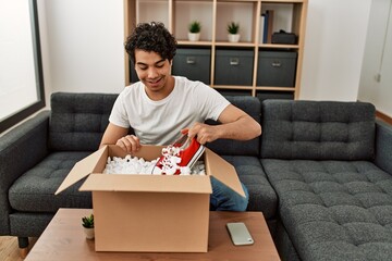 Wall Mural - Young hispanic man unboxing sneakers of cardboard box sitting on the sofa at home.