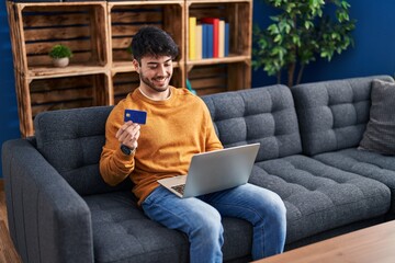 Wall Mural - Young hispanic man using laptop and credit card sitting on sofa at home