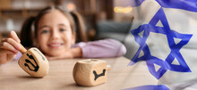Banner With Israeli National Flag And Happy Girl With Dreidels Celebrating Hannukah At Home