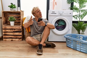 Canvas Print - Young blond man doing laundry using smartphone pointing fingers to camera with happy and funny face. good energy and vibes.