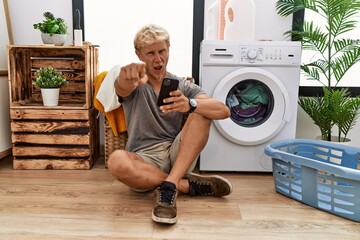 Sticker - Young blond man doing laundry using smartphone pointing displeased and frustrated to the camera, angry and furious with you