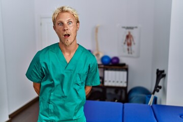Wall Mural - Young blond man wearing physiotherapist uniform standing at clinic afraid and shocked with surprise expression, fear and excited face.