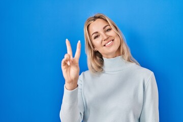 Poster - Young caucasian woman standing over blue background smiling looking to the camera showing fingers doing victory sign. number two.