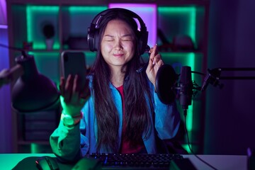 Wall Mural - Young asian woman playing video games with smartphone gesturing finger crossed smiling with hope and eyes closed. luck and superstitious concept.