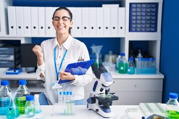 Sticker - Young brunette woman working at scientist laboratory very happy and excited doing winner gesture with arms raised, smiling and screaming for success. celebration concept.