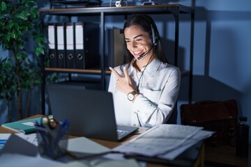 Wall Mural - Young brunette woman wearing call center agent headset working late at night cheerful with a smile of face pointing with hand and finger up to the side with happy and natural expression on face