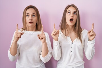 Canvas Print - Middle age mother and young daughter standing over pink background amazed and surprised looking up and pointing with fingers and raised arms.