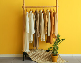 Wall Mural - Rack with trendy clothes, rug and houseplant near yellow wall