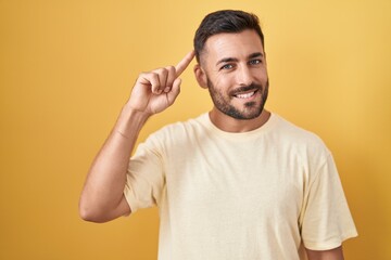 Wall Mural - Handsome hispanic man standing over yellow background smiling pointing to head with one finger, great idea or thought, good memory
