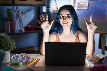 Poster - Young modern girl with blue hair sitting at art studio with laptop at night showing and pointing up with fingers number seven while smiling confident and happy.