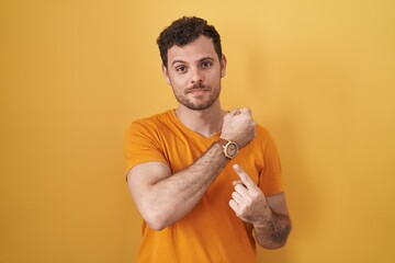 Canvas Print - Young hispanic man standing over yellow background in hurry pointing to watch time, impatience, looking at the camera with relaxed expression