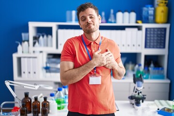 Wall Mural - Young hispanic man working at scientist laboratory smiling with hands on chest with closed eyes and grateful gesture on face. health concept.