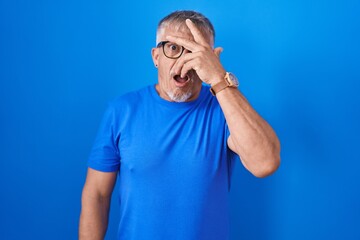 Canvas Print - Hispanic man with grey hair standing over blue background peeking in shock covering face and eyes with hand, looking through fingers with embarrassed expression.