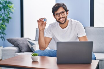 Canvas Print - Handsome latin man holding virtual currency bitcoin using laptop looking positive and happy standing and smiling with a confident smile showing teeth