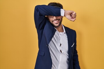 Poster - Handsome latin man standing over yellow background covering eyes with arm smiling cheerful and funny. blind concept.