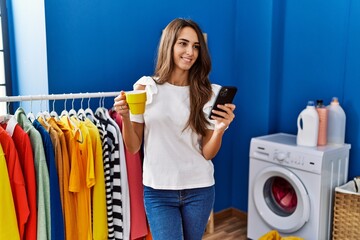 Wall Mural - Young hispanic woman drinking coffee and using smartphone at laundry room