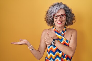 Middle age woman with grey hair standing over yellow background showing palm hand and doing ok gesture with thumbs up, smiling happy and cheerful