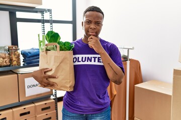 Canvas Print - Young african volunteer man holding groceries serious face thinking about question with hand on chin, thoughtful about confusing idea