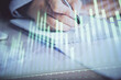 © peshkova - A woman hands writing information about stock market in notepad. Forex chart holograms in front. Concept of research. Multi exposure
