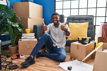 Wall Mural - African american man sitting on the floor at new home success sign doing positive gesture with hand, thumbs up smiling and happy. cheerful expression and winner gesture.