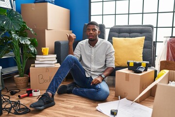 African american man sitting on the floor at new home surprised pointing with hand finger to the side, open mouth amazed expression.