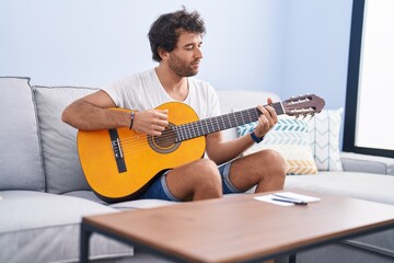 Wall Mural - Young hispanic man playing classical guitar sitting on sofa at home