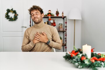 Poster - Young handsome man with beard sitting on the table by christmas decoration smiling with hands on chest with closed eyes and grateful gesture on face. health concept.