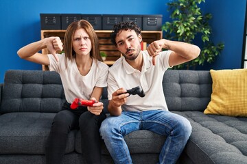 Poster - Young couple playing video game holding controller at home with angry face, negative sign showing dislike with thumbs down, rejection concept