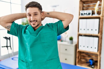 Poster - Young physiotherapist man working at pain recovery clinic relaxing and stretching, arms and hands behind head and neck smiling happy