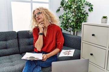 Wall Mural - Middle age psychologist woman at consultation office with hand on chin thinking about question, pensive expression. smiling with thoughtful face. doubt concept.