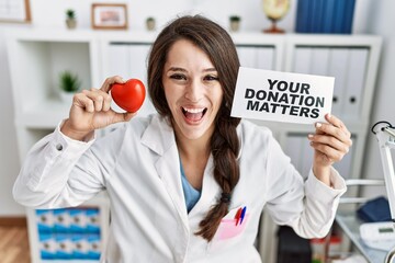 Wall Mural - Young doctor woman holding your donation matters banner at the clinic smiling and laughing hard out loud because funny crazy joke.